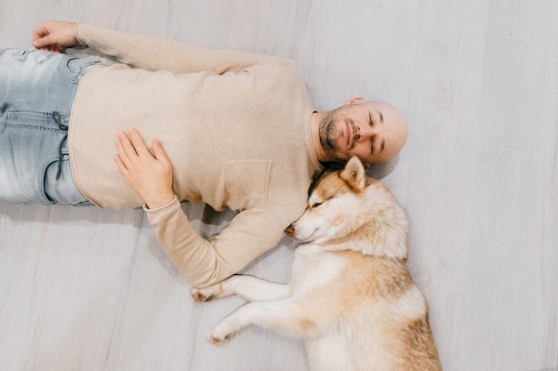 Adult bald man with husky puppy sleeping on floor. Owner with pet together at home. Lovely dog resting with young male.