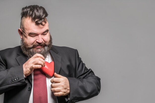 Foto uomo barbuto adulto in una giacca nera che tiene un cuore rosso sfondo grigio