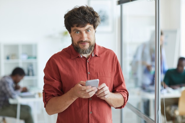 adult bearded man dressed in casual wear and holding smartphone while standing in office