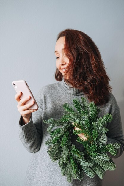 Adult beautiful smiling woman forty years with brunette curly hair in warm grey knitted dress with diy fir christmas wreath in hand using mobile phone on grey background