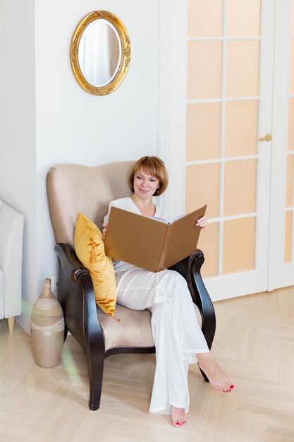 大人の美しい女性がスタイリッシュなリビング ルームで時間を過ごし、大きな本を手に持っています。