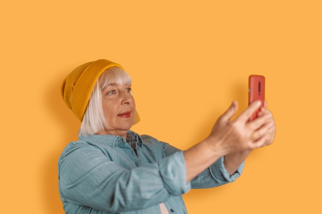 Adult beautiful woman in stylish clothes taking a selfie posing isolated on yellow orange background