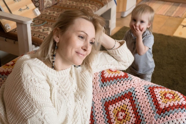 Фото Взрослая блондинка сидит дома на диване, расслабляясь, пока маленькая дочь играет одна