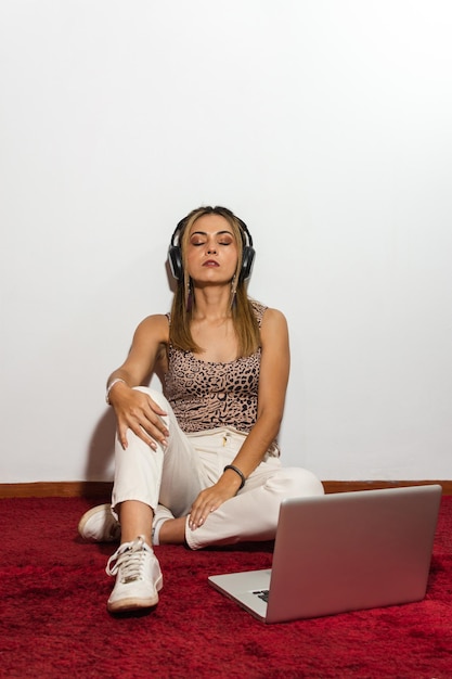 Взрослая блондинка расслабляется, используя наушники и ноутбук, слушает музыку, сидя на красной ковровой дорожке и на белом фоне