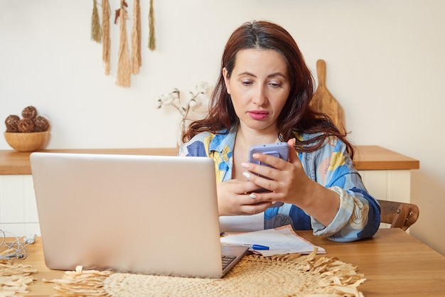 An adult brunette uses a mobile phone works on a laptop The concept of remote work learning