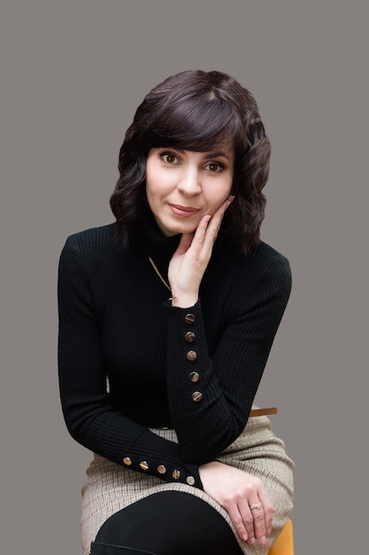 Adult brunette woman in gray skirt sits on gray background.