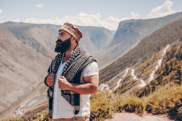 An adult brutal bearded man in a hat and a patterned scarf looks mysteriously to the side against the background of a vast rocky mountain landscape. Admire the view.