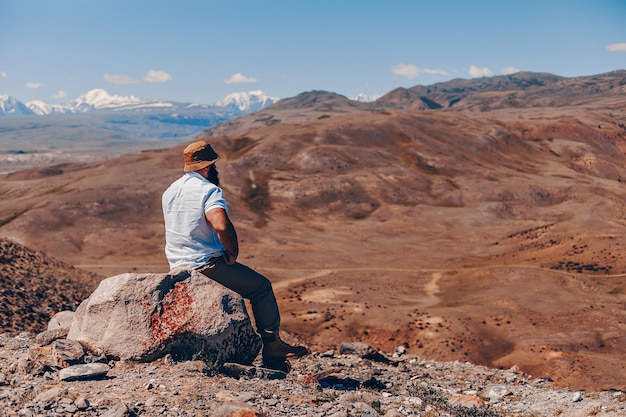 Un uomo adulto brutale con la barba con un cappello panama e una camicia bianca si siede su un ciottolo con la schiena e guarda il paesaggio come se fosse con montagne pittoresche.