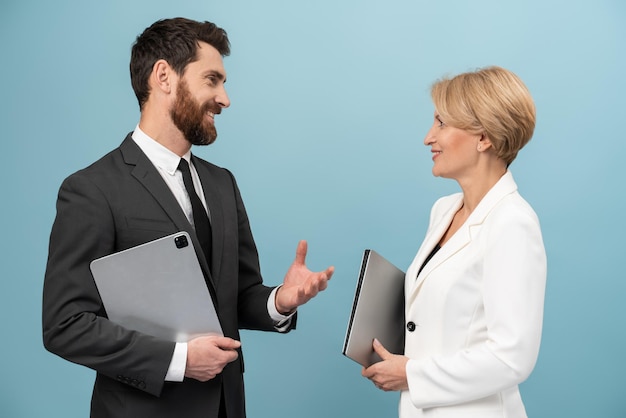 Adult businessman and senior businesswoman wearing formal suits holding laptops and chatting with each other Isolated over blue background