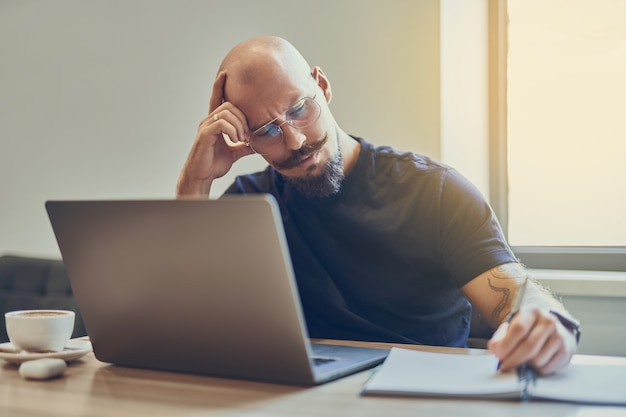 Adult caucasian bald man manager using laptop while writing notes in notebook at workplace