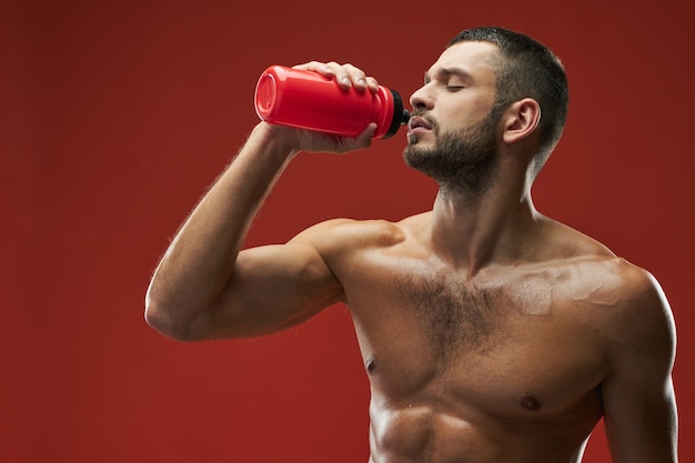 Фото Взрослый кавказский бородатый спортсмен держит красную бутылку с напитком на красном фоне