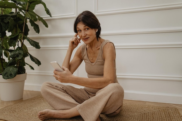 Adult caucasian brunette woman with short hair sits on floor in living room browsing phone chatting online with friends