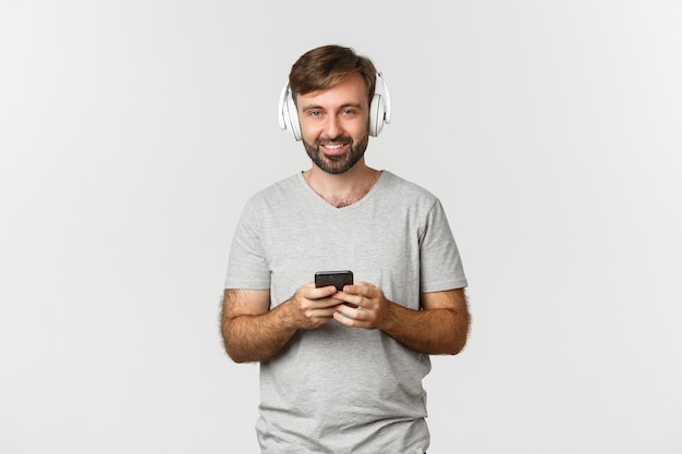 Взрослый кавказский парень улыбается, слушает музыку в наушниках и использует мобильный телефон, стоя над
