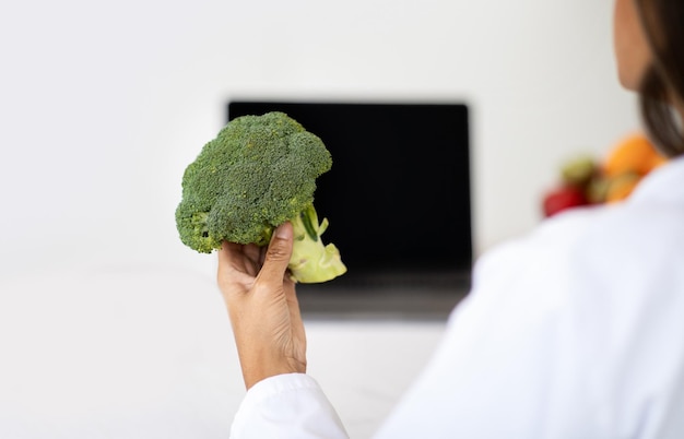 Adult caucasian nutritionist doctor in white coat at table shows broccoli in laptop with blank