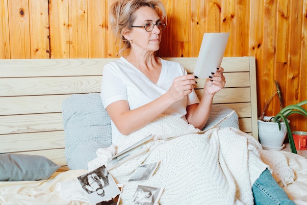 Adult caucasian woman nostalgic for youth and childhood looking at old family album photos while relaxing in bed in bedroom. Middle aged woman in glasses holding vintage photographs reminiscing