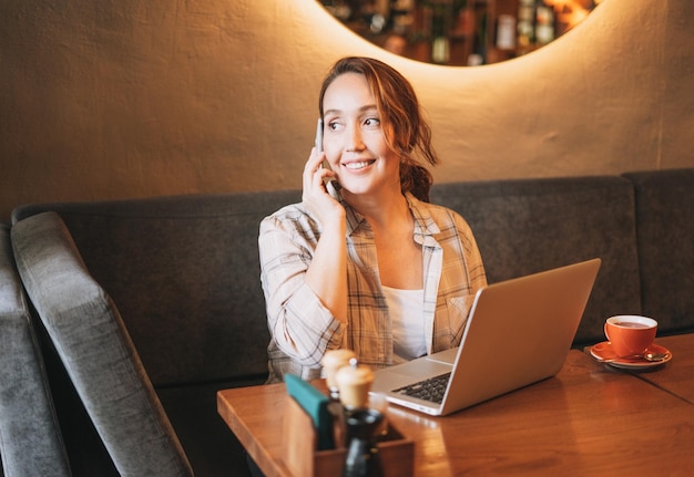 Adult charming brunette woman in plaid shirt working with laptop using mobile phone at the cafe