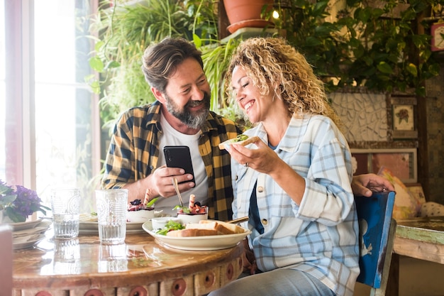 성인 커플은 웃고 점심과 전화 화상 통화를 즐기고 바에 앉아 행복과 함께 웃고 있습니다. 남자와 여자는 셀룰러를 사용하고 레스토랑에서 브런치를 먹습니다.