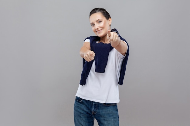 Adult cute woman in casual clothes choice fingers at camera with jeans and sweater on her shoulders, smart watch on arm, Studio shot, isolated on grey background