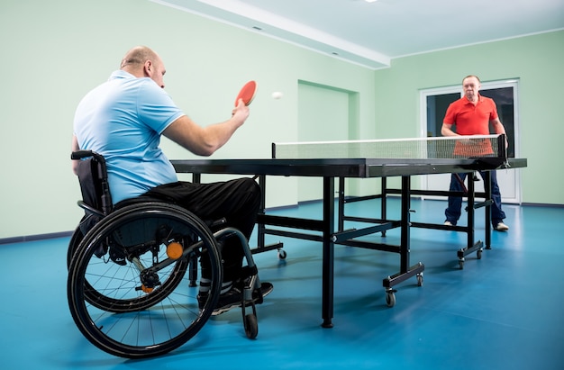 Adult disabled man in a wheelchair play at table tennis with his coach