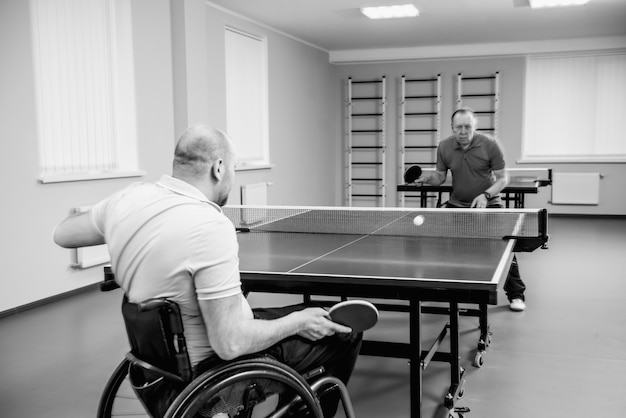 Взрослый человек-инвалид в инвалидной коляске играет в настольный теннис со своим тренером