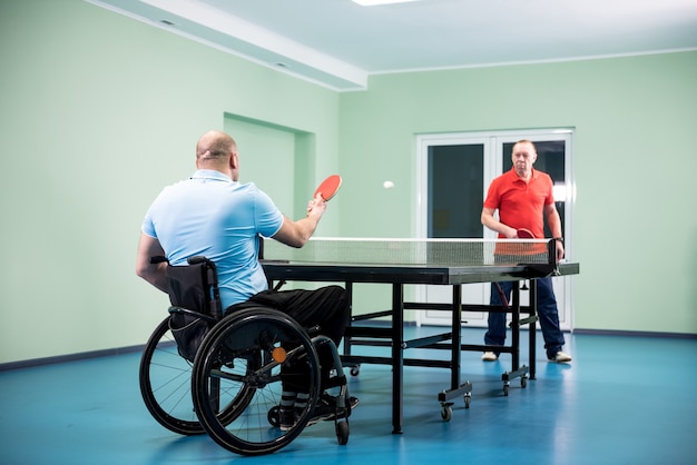 Adult disabled man in a wheelchair play at table tennis with his coach