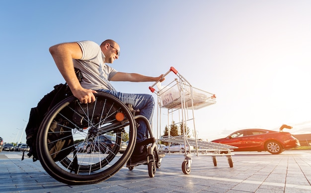 Adult disabled man in a wheelchair pushes a cart towards a car in a supermarket parking lot