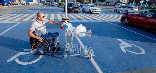 Adult disabled man in a wheelchair pushes a cart towards a car in a supermarket parking lot