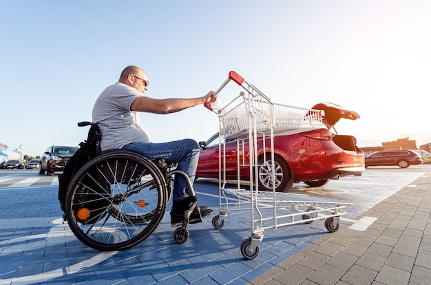 Adult disabled man in a wheelchair pushes a cart towards a car in a supermarket parking lot