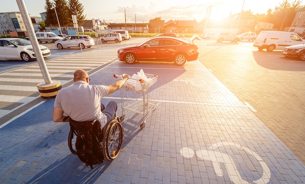 Adult disabled man in a wheelchair pushes a cart towards a car in a supermarket parking lot