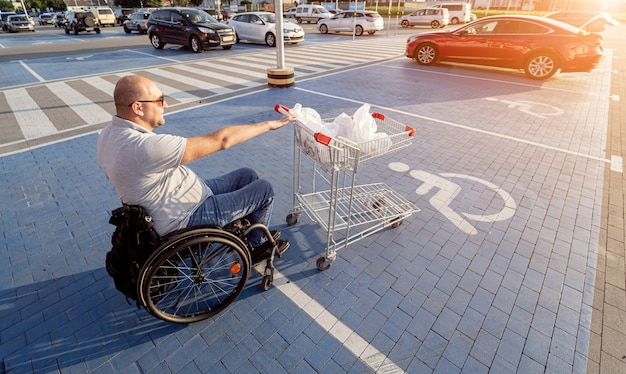 Adult disabled man in a wheelchair pushes a cart towards a car in a supermarket parking lot