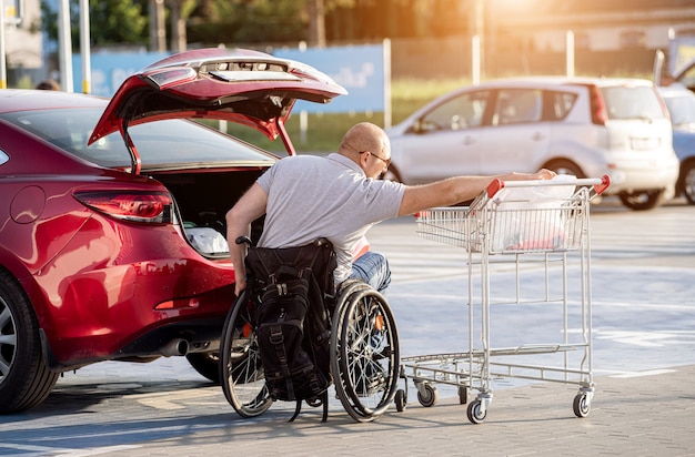 Adult disabled man in a wheelchair puts purchases in the trunk of a car in a supermarket parking lot