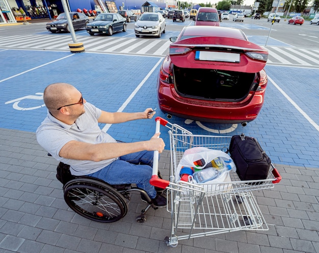 Adult disabled man in a wheelchair puts purchases in the trunk of a car in a supermarket parking lot