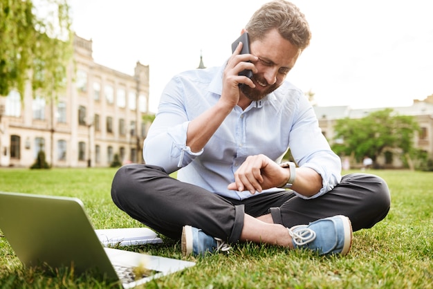 adult european man in business clothing, looking at smartwatch while sitting on grass in park with legs crossed and having business call