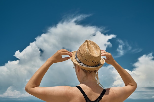 Adult european woman is on the seashore and holds a straw hat\
on her head with her hands a silhouette of a beautiful blonde\
against the background of cumulus clouds over the sea