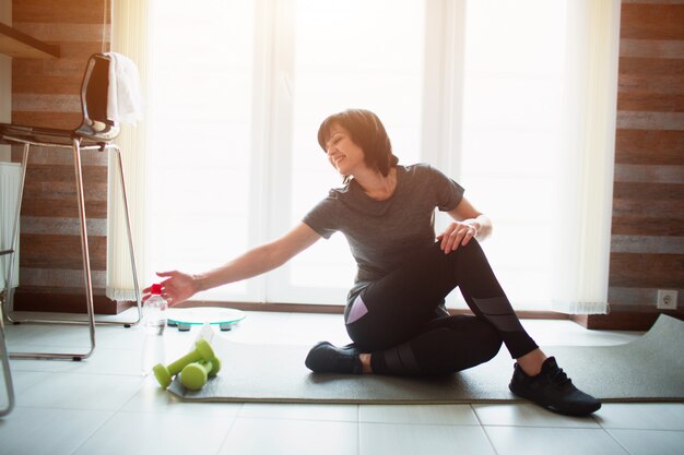 Adult fit slim woman has workout at home. Senior female model reaching water bottle with smile. Sit on yoga mat during exercise break. Training alone in room.