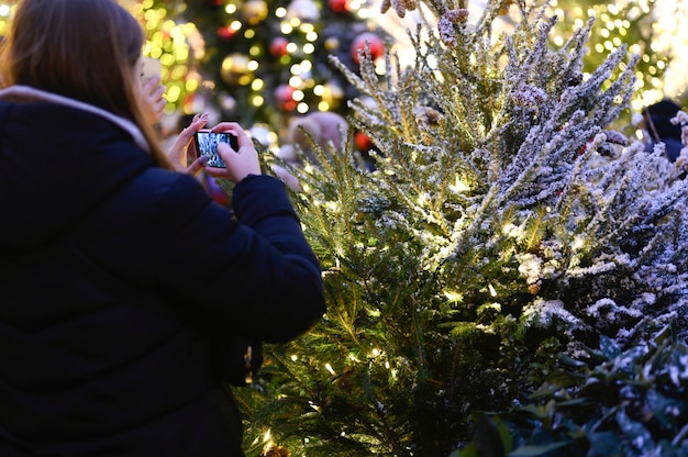 성인 소녀는 밤, 후면보기에 빛나는 불빛으로 화환으로 장식 된 크리스마스 트리의 그녀의 전화로 사진을 찍습니다.
