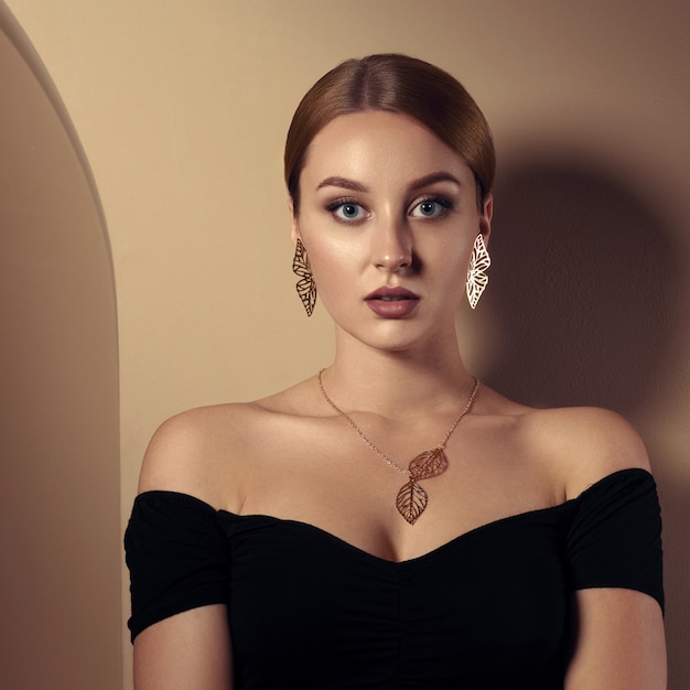 Adult girl wearing nature inspired golden jewelry set and black dress and leaning in beige room