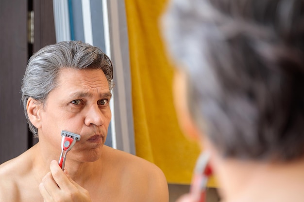 Взрослый седоволосый мужчина брит бритвой перед зеркалом