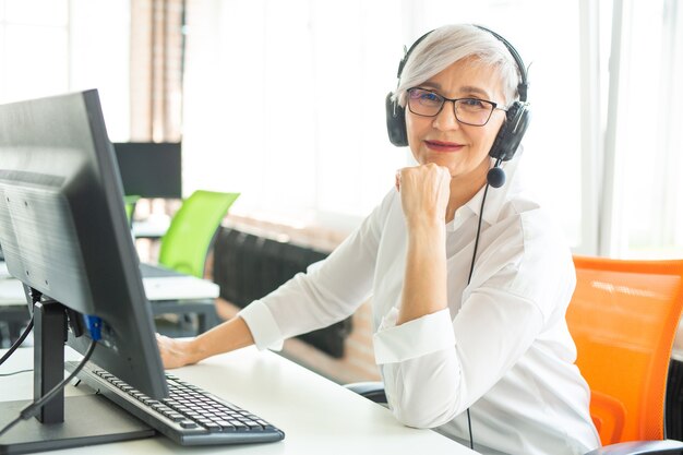 adult grayhaired woman at the age of working in the office at the computer in headphones with a microphone