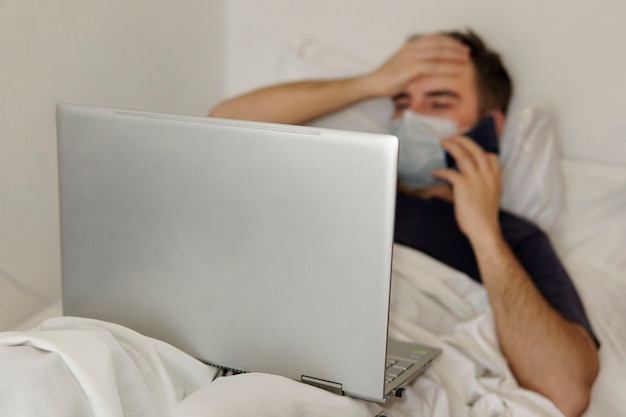 An adult guy is lying in a hospital bed with a laptop holding his head. Concept of remote work during the coronavirus pandemic