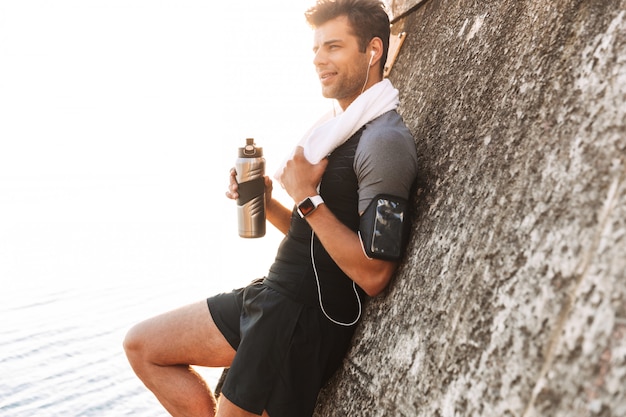 Взрослый парень в спортивном костюме слушает музыку через беспроводные наушники и пьет воду из металлической кружки