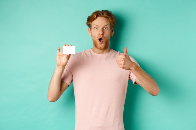 Взрослый парень с рыжими волосами и бородой показывает пластиковую кредитную карту и большой палец вверх, выглядит впечатленным, рекомендует банк, стоя на фоне мяты.