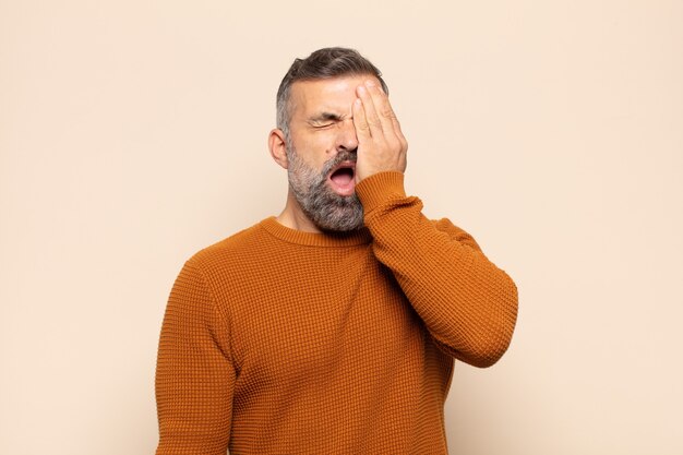 Adult handsome man looking sleepy, bored and yawning, with a headache and one hand covering half the face