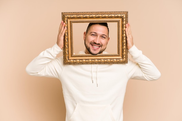 Adult latin man holding a vintage frame and smiling isolated