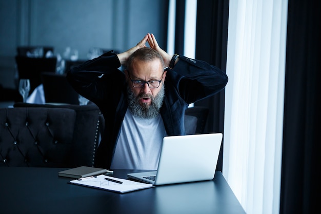 An adult male businessman is working on a new project and looking at the stock growth charts. Sits at the table by the large window. Looks at the laptop screen and smiles