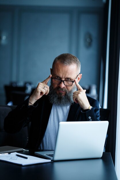 An adult male businessman is working on a new project and looking at the stock growth charts. Sits at the table by the large window. Looks at laptop screen