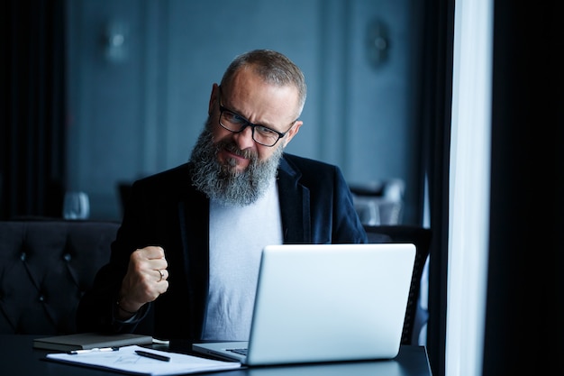 An adult male businessman is working on a new project and looking at the stock growth charts. Sits at the table by the large window. Looks at laptop screen