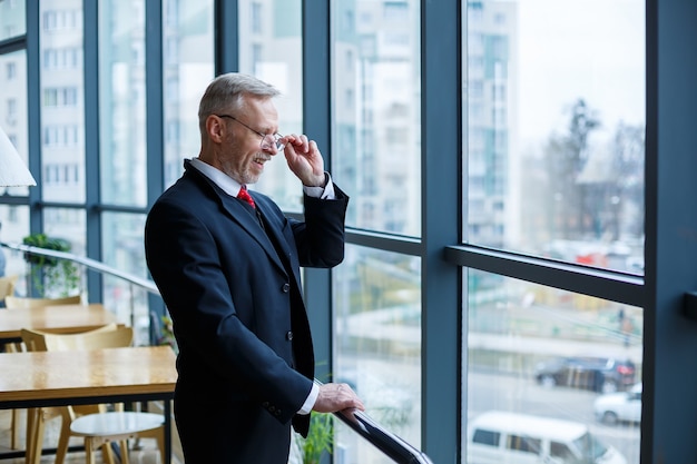 Adult male businessman, teacher, mentor looks out the large window of his office. Work in the office. Development of a new business project