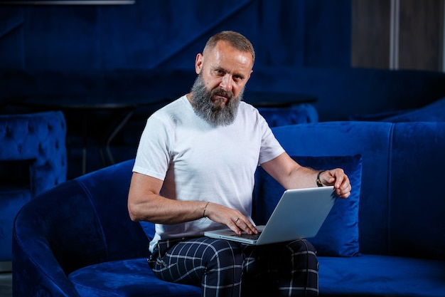 Adult male businessman, teacher, mentor working on a new project. Sits by a large window on the table. He looks at the laptop screen.