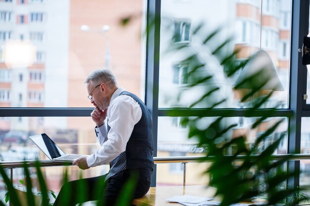 Adult male businessman, teacher, mentor working on a new project. Sits by a large window on the table. He looks at the laptop screen.